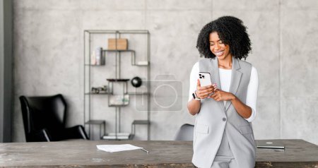 Diese afroamerikanische Geschäftsfrau strahlt Positivität aus und lächelt, während sie ihr Smartphone benutzt, das in einer schnelllebigen technologischen Welt eindeutig floriert. Ihre professionelle Kleidung unterstreicht den Geschäftssinn