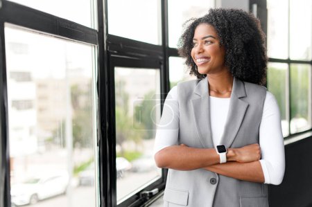 Eine optimistische afroamerikanische Geschäftsfrau blickt aus dem Fenster, ihr nachdenklicher Ausdruck und ihre entspannte Haltung vermitteln strategisches Denken und Zukunftsplanung in einem hellen Büro.