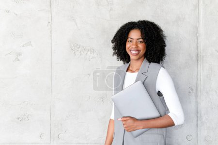 Téléchargez les photos : Une femme d'affaires afro-américaine se tient confiante avec son ordinateur portable, son sourire exsudant un professionnalisme dans un contexte minimaliste. Cette image reflète l'essence du succès moderne de l'entreprise - en image libre de droit