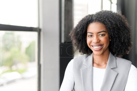 A radiant African-American businesswoman smiles warmly in an office setting, showcasing a blend of professionalism and welcoming charisma that represents approachable leadership.