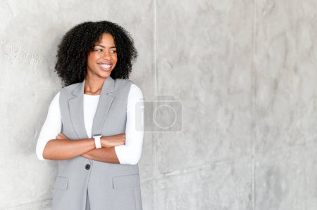 Die Geschäftsfrau steht selbstbewusst mit den Armen an ihrer Seite, ein subtiles Lächeln spielt auf ihren Lippen und verkörpert Stärke und Nahbarkeit in ihrem grauen Ensemble vor der strukturierten Wand.