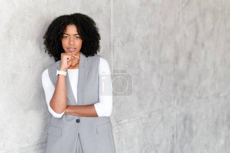 Una mujer de negocios afroamericana posa pensativamente, con la barbilla apoyada en su mano, sobre un fondo gris texturizado, simbolizando la contemplación y el profesionalismo en un entorno corporativo.