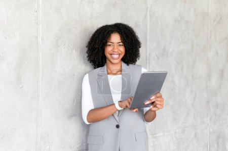 An exuberant African-American businesswoman in professional attire holds a digital tablet, her radiant smile and engaging eye contact suggesting approachability and tech-savviness in corporate setting