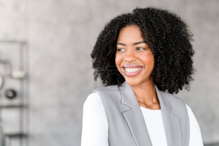Foto de Una radiante empresaria afroamericana sonríe con confianza, en un ambiente de oficina moderno. Su atuendo profesional y su actitud alegre proyectan tanto la accesibilidad como la autoridad. - Imagen libre de derechos