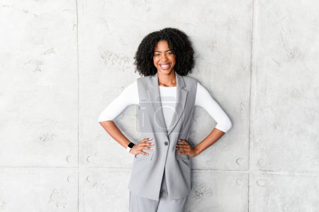 Photo for A poised African-American businesswoman stands with hands on her hips, wearing a stylish grey sleeveless vest and trousers, radiating confidence and professionalism against a textured grey background. - Royalty Free Image