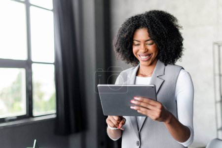 Téléchargez les photos : Une femme d'affaires afro-américaine en costume gris sourit en s'engageant avec sa tablette, debout dans un bureau. Ce plan met l'accent sur l'activité commerciale dynamique et l'intégration transparente de la technologie - en image libre de droit