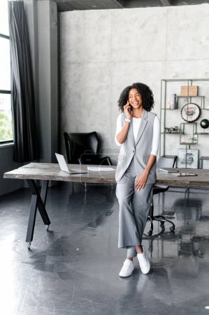 A smiling African-American businesswoman leans on a table during a phone call, exuding confidence and comfort in her professional realm. Full length