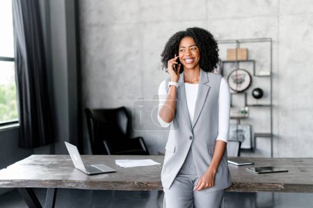 A joyful African-American businesswoman engages in a phone call, her expression one of satisfaction and command, set against the backdrop of an industrial-chic office space