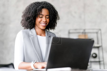 An African-American businesswoman works on her laptop, smiling and radiating satisfaction, embodying the success and enjoyment of her professional endeavors.