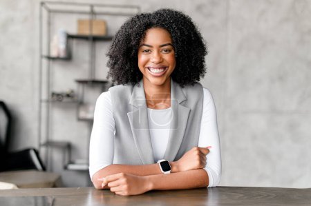 Eine charismatische afroamerikanische Geschäftsfrau grüßt mit einem warmen Lächeln von ihrem Schreibtisch und verkörpert Professionalität und eine einladende Arbeitsatmosphäre in einem modernen Ambiente..