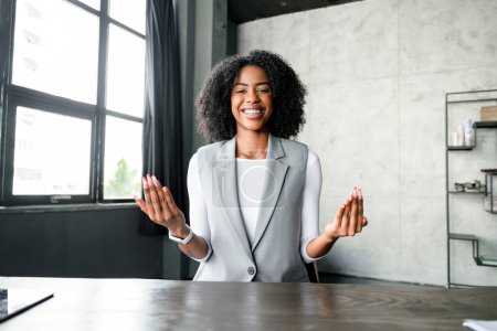 Una mujer de negocios afroamericana sonríe alegremente mientras hace gestos durante una videoconferencia, su estilo de comunicación carismático realza su mensaje en un entorno de oficina moderno y minimalista..