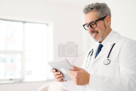 A senior doctor with a gentle smile reviews data on a tablet, a sign of embracing modern medical technology in patient care, side view. Healthcare and technology concept