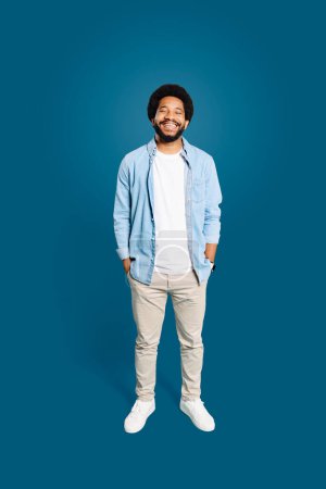 Ein fröhlicher Mann mit natürlicher Afrofrisur steht selbstbewusst da, die Hände in den Taschen, in einem lässigen Jeanshemd und einem echten Lächeln vor tiefblauem Hintergrund, in voller Länge.