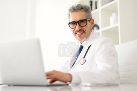 Senior doctor with grey hair looks at camera while using a laptop, suggesting adaptability to modern medical technology, friendly expression and attentive posture convey a commitment to patient care