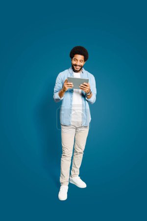 Hombre brasileño mira en la pantalla de su tableta, sonriendo, ya que sostiene el dispositivo de una manera relajada, indicativo del uso cómodo e intuitivo de los aparatos de tecnología moderna, longitud completa aislada en azul