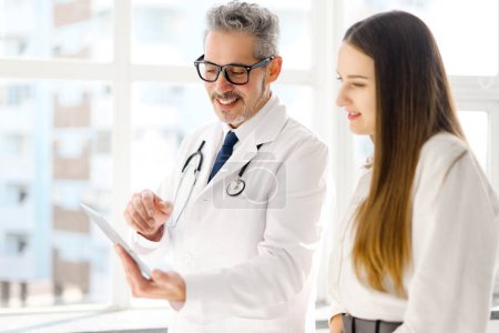 Ein angesehener Arzt mit silbernem Haar konsultiert zusammen mit einer jungen Frau ein Tablet und betont einen interaktiven und technisch versierten Ansatz im Gesundheitswesen.