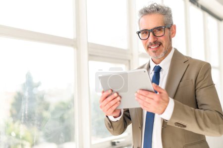 Photo for A senior businessman engages with modern technology, holding a tablet with an assured smile, showcasing how traditional business acumen blends with contemporary tools - Royalty Free Image