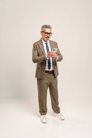 A senior businessman engages with a smartphone in a focused manner, symbolizing the fusion of experience and modern technology in the business realm. Full-length portrait