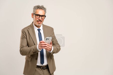 Foto de El hombre de negocios maduro de pelo gris sonríe y mira a la cámara mientras usa su teléfono inteligente, mostrando a un profesional senior navegando cómodamente por la tecnología de comunicación moderna - Imagen libre de derechos