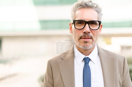 A senior businessman in glasses with grey hair poses confidently outdoors, his sharp beige suit and blue tie signaling a polished and authoritative presence. The concept of leadership and expertise