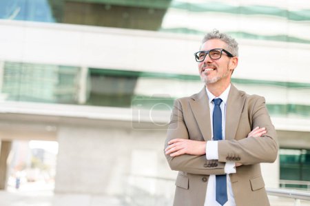 Photo for Senior businessman stands confidently with arms crossed in front of a modern office building, his expression content and optimistic, embodying success and experience. Leadership and career achievement - Royalty Free Image