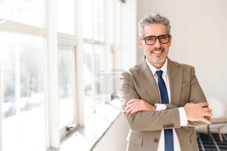 A senior businessman in a chic beige suit and navy blue tie stands with arms confidently folded in the bright office. The experienced and positive professional person indoor