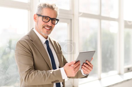 The senior executive, standing in a naturally lit room with a large window view, navigates through a tablet with ease, showcasing the seamless integration of technology in modern corporate life.