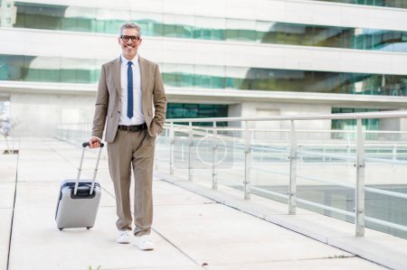 Ein charismatischer Geschäftsmann mit einem Koffer posiert selbstbewusst im Freien, sein Lächeln deutet auf eine positive Perspektive und die Bereitschaft für neue Geschäftsmöglichkeiten hin. Reise- und Geschäftskonzept