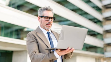 Senior businessman reviews content on laptop outdoors, focused and absorbed in his work, with an office building as his backdrop, professional stays connected and productive in various environments