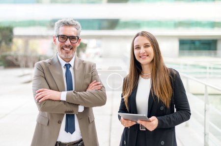 Un alto ejecutivo equilibrado con el pelo gris se levanta con confianza con los brazos cruzados, acompañado por una profesional que sostiene una tableta, simbolizando liderazgo y experiencia.