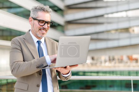 L'homme d'affaires expérimenté est capturé à mi-geste, interagissant avec son écran d'ordinateur portable, dans le contexte d'un immeuble de bureaux en verre, dépeignant un engagement actif avec la technologie
