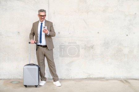 Der Geschäftsmann steht mit seinem Smartphone an seinem Koffer vor einem schlichten Hintergrund, der die Integration von Technologie und Mobilität im Geschäftsleben symbolisiert. Reise- und Geschäftskonzept