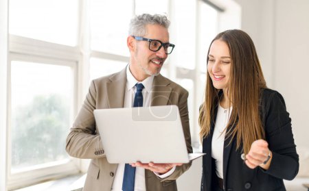 Senior businessman with grey hair shares a light-hearted moment with a female colleague while working on a laptop, their easy camaraderie evident in a modern office in a positive workplace atmosphere