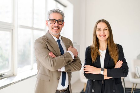 Photo for A charismatic senior entrepreneur with grey hair stands arms crossed next to a young professional woman, both smiling confidently in a modern office, reflecting a successful business partnership - Royalty Free Image