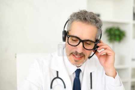 Un médico senior está totalmente involucrado en una llamada de telesalud, usando un auricular y una computadora portátil, mostrando cómo se usa la tecnología moderna en las consultas de los pacientes y la comunicación de la atención médica..