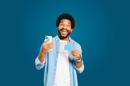 Voller Aufregung blickt der junge Brasilianer in die Kamera, während er nach einer erfolgreichen Online-Transaktion eine Kreditkarte und ein Smartphone in der Hand hält oder, isoliert auf blauem Grund, gute Finanznachrichten erhält.