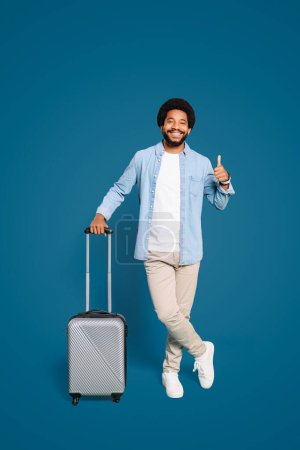 A cheerful young Brazilian man with a captivating smile holds a silver suitcase, giving a thumbs-up against a vibrant blue background, exuding a sense of excitement and anticipation for travel