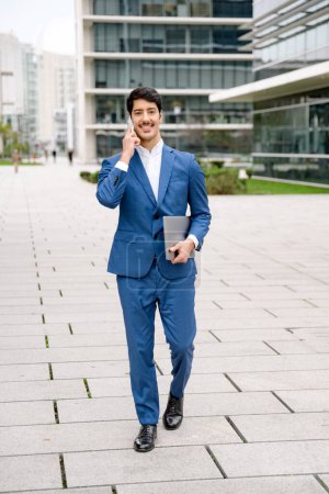 Téléchargez les photos : Dans un paysage urbain animé, un jeune homme d'affaires hispanique sourit en faisant un appel d'affaires, avec un ordinateur portable à la main, symbolisant l'agilité et la connectivité des professionnels modernes. - en image libre de droit