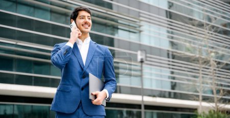 Photo for Confident smart Hispanic businessman in a blue suit happily converses on his smartphone while holding a laptop, showcasing flexibility and connectivity in the corporate world. - Royalty Free Image