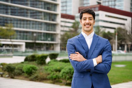 Hispanic businessman is pictured with a bright smile, standing arms crossed in a power pose that suggests confidence and readiness for business challenges ahead, set against a blurred urban setting.