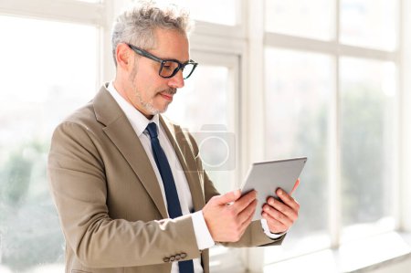 Un homme d'affaires distingué dans un costume beige sur mesure opère en toute confiance une tablette, reflétant l'intégration dynamique de la technologie dans les affaires, soulignant la fusion de l'expérience et de l'innovation