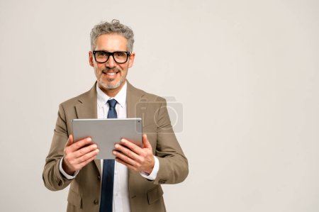 A cheerful senior businessman confidently stands with a tablet in hands, indicating the importance of digital devices in the business landscape and executive accessibility.