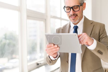 Ein hochrangiger Geschäftsmann im beigen Anzug steht vor einem hellen Fenster und lächelt, während er mit einem Tablet hantiert. Dieses Bild fängt das Konzept der modernen Technologie in den Händen erfahrener Fachleute ein.