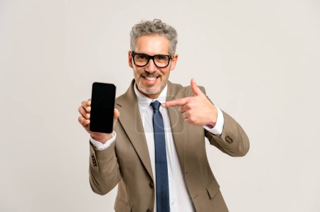 With a charismatic smile, a grey-haired businessman points to a smartphone with an empty screen, suggesting a place for advertising, copy space. Man in suit paying attention on pone display, mock up
