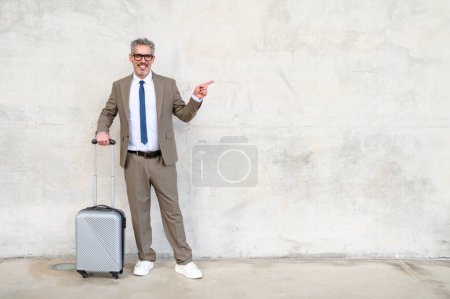 Un hombre de negocios animado con el pelo gris apunta hacia un lado mientras está de pie con su maleta de viaje, tal vez indicando direcciones u oportunidades, contra una pared de hormigón minimalista.