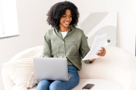 Femme afro-américaine joyeuse s'assoit sur un canapé avec ordinateur portable, regardant à travers les documents, son visage exprimant sa satisfaction dans un cadre accueillant et contemporain. Étudiante se préparant aux examens de la maison