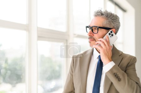 The grey-haired businessman is speaking on his smartphone with a look of concentration, in a bright office space. This image portrays the dynamic nature of business communication