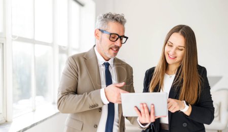 A male entrepreneur with grey hair and a welcoming smile holds a tablet, while a young female professional looks on with interest in a bright office. Collaborative interaction in a modern business