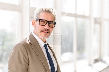 Un homme d'affaires mûr aux cheveux gris et au sourire chaleureux se tient confiant dans un bureau bien éclairé, son costume pointu et ses lunettes parlant de sagesse et de professionnalisme moderne.