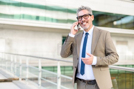 Ein hochrangiger Profi in schicker Businesskleidung führt ein angenehmes Telefongespräch und gestikuliert mit einer Hand, während er mit dem modernen Bürogebäude im Hintergrund spricht.